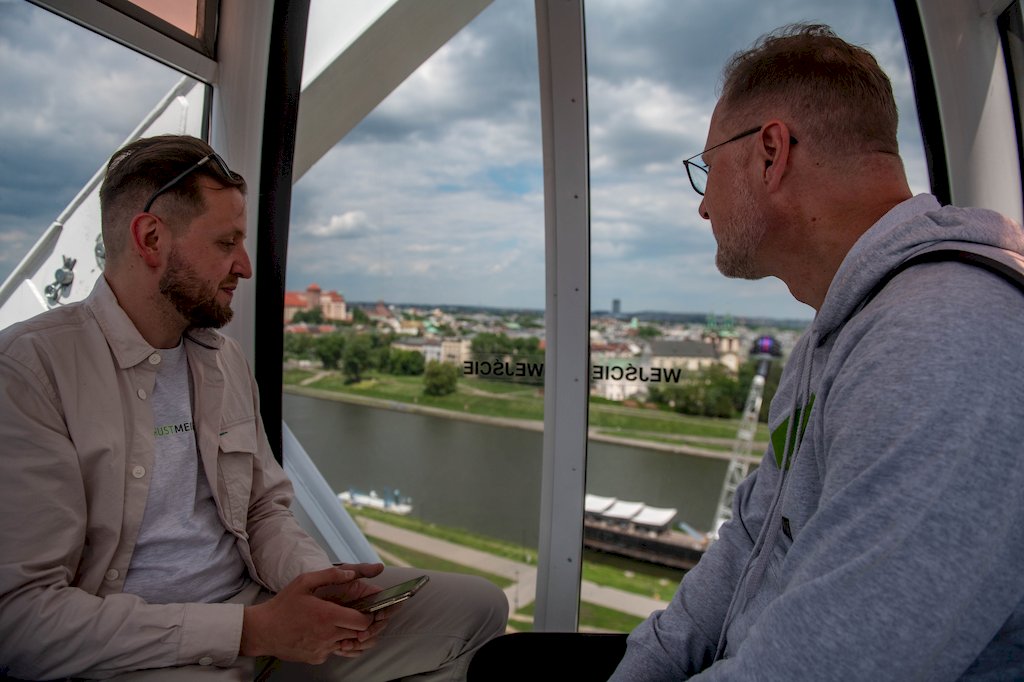 Dwóch mężczyzn siedzących naprzeciwko siebie patrzy z przeszklonej kabiny wagonika karuzeli na Wisłę i panoramę Krakowa