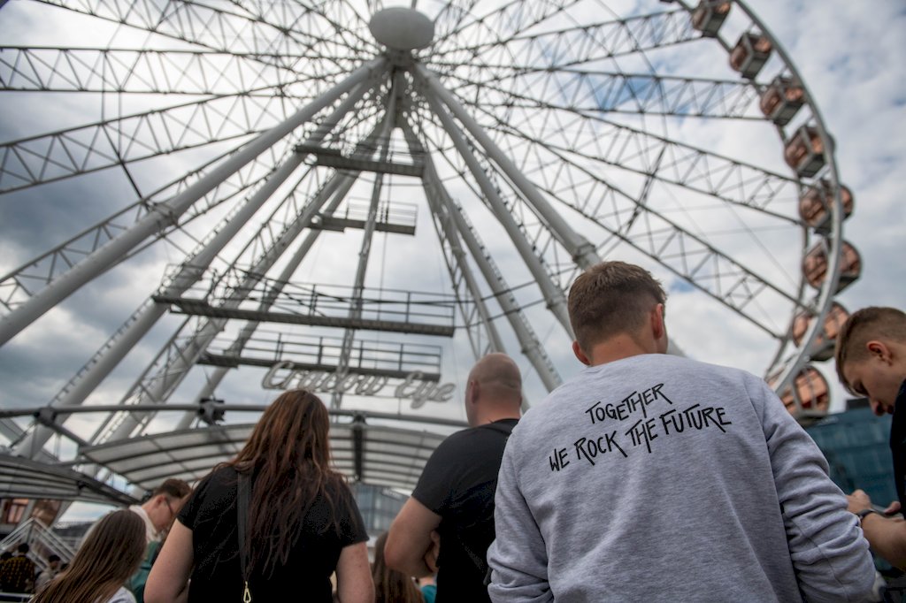Kilka osób stojących tyłem na tle karuzeli Cracow Eye. Jedna w szarej bluzie z napisem Together we rock the future