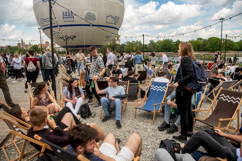 Studenci rozmawiają siedząc na leżakach lub stojąc. W tle balon, Wisła i na horyzoncie Wawel.