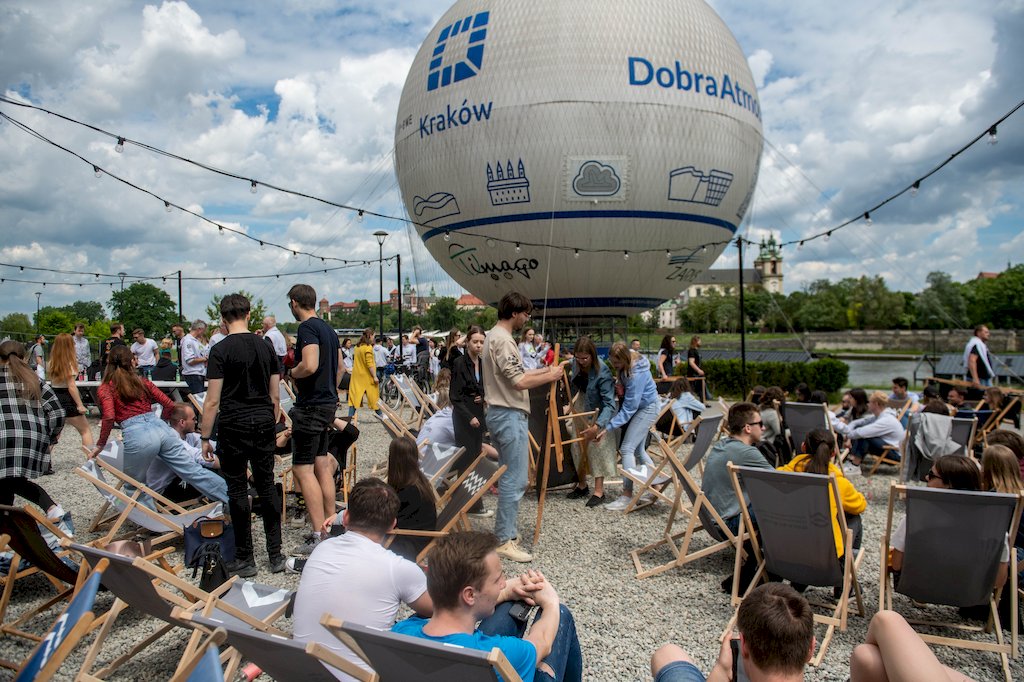 Studenci i studentki siedzą na leżakach i rozmawiają. W tle balon z napisem Kraków i Dobra atmosfera