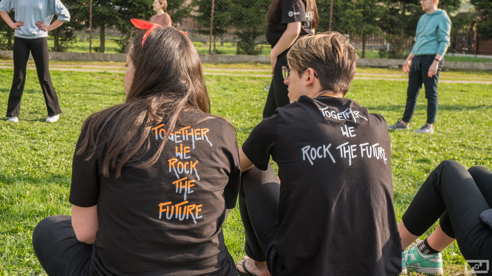 Studentka i student siedzą na trawie obok siebie. Ubrani są w czarne T-Shirty z hasłem: Together we rock the future.