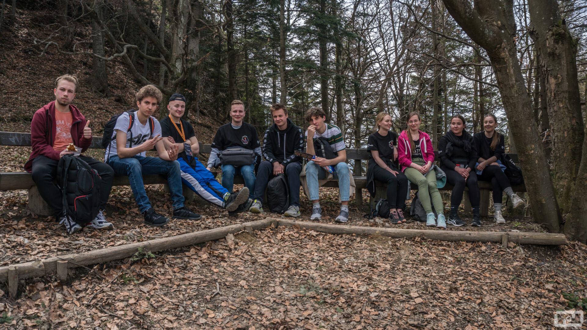 Studenci i studentki ubrani w stroje górskie siedza na drewnianej ławce w lesie. Przed nimi znajdują się opadające liście..