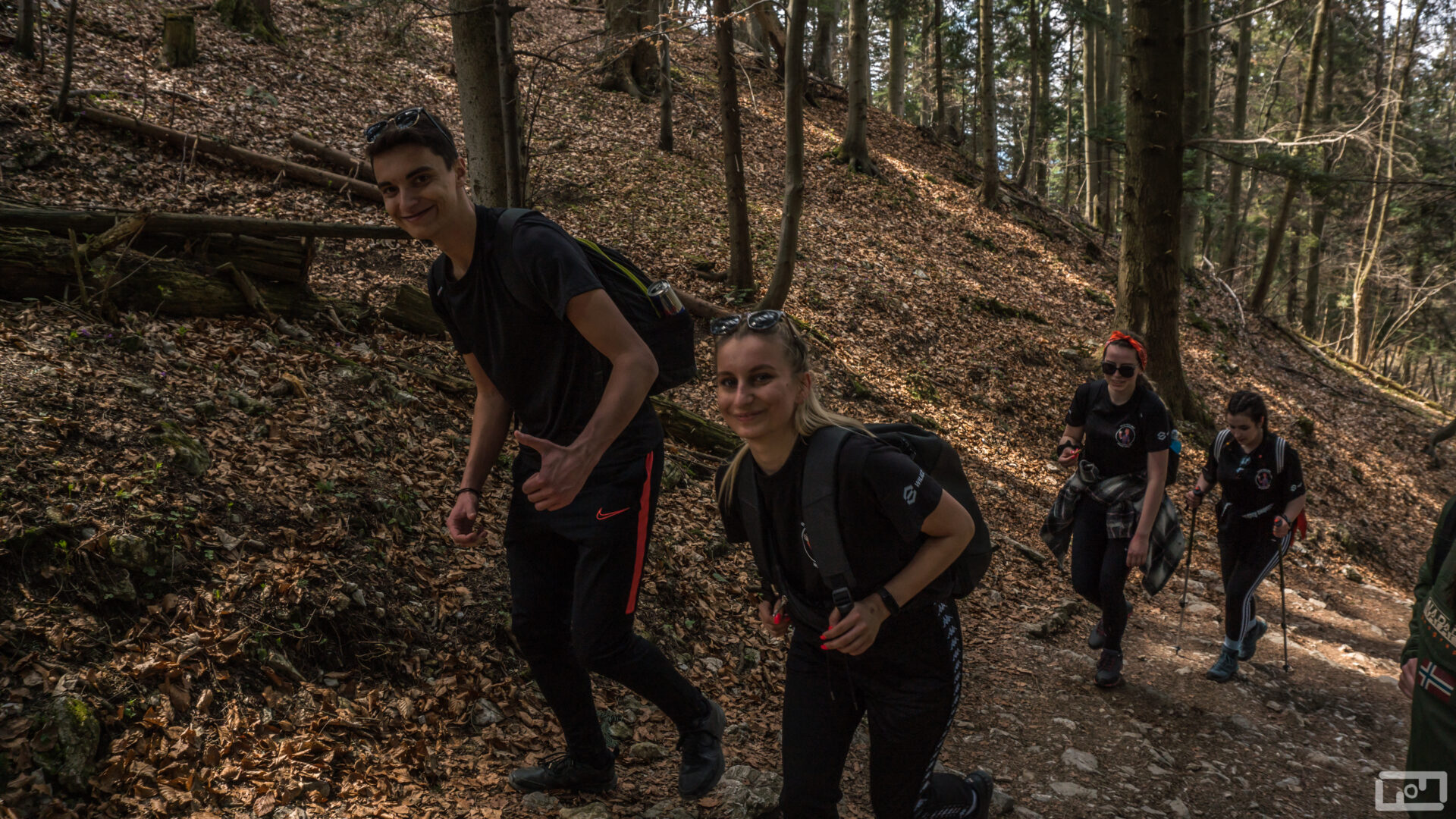 Studenci i studentki ubrani w stroje górskie z założonymi plecakami idą szlakiem przez las.