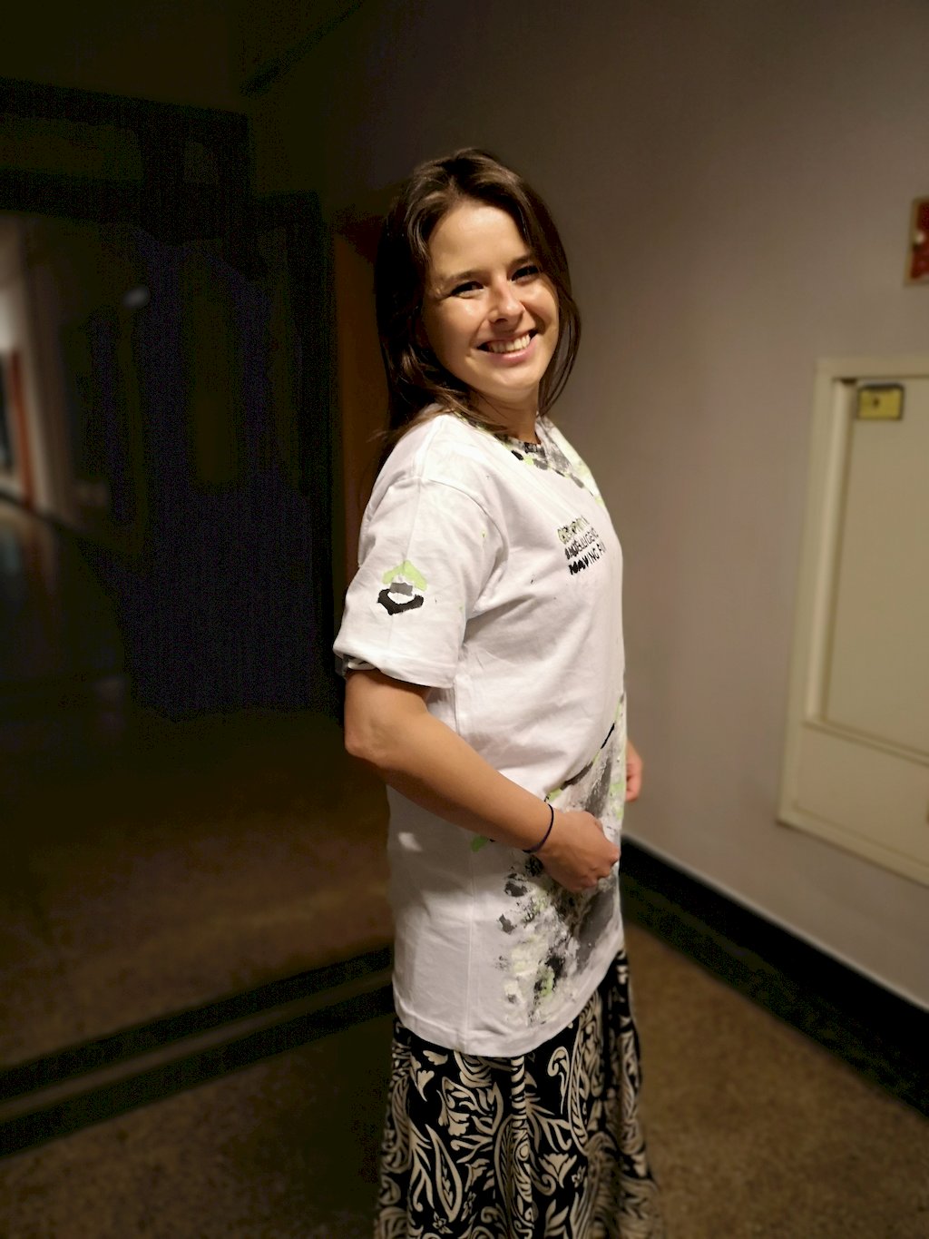 Młoda kobieta stojąca na korytarzu budynku prezentuje koszulke z wymalowanym na krótkim rękawku logo WILIGZ.
