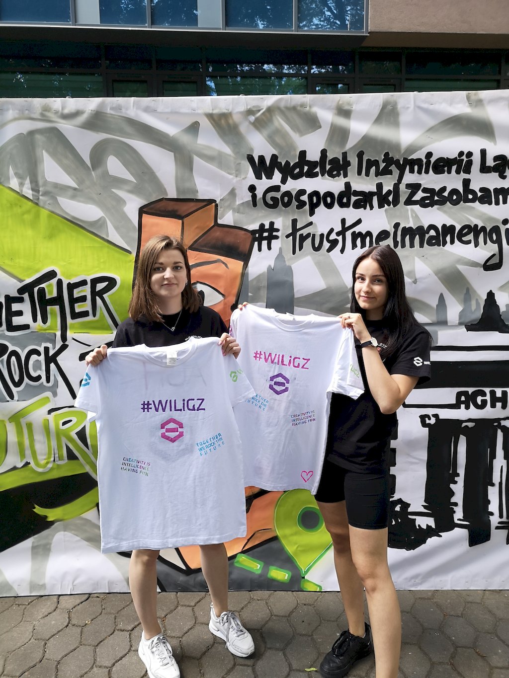 Dwie studentki prezentuja koszulki z namalowanym sygnetem logo i napisem WILiGZ.