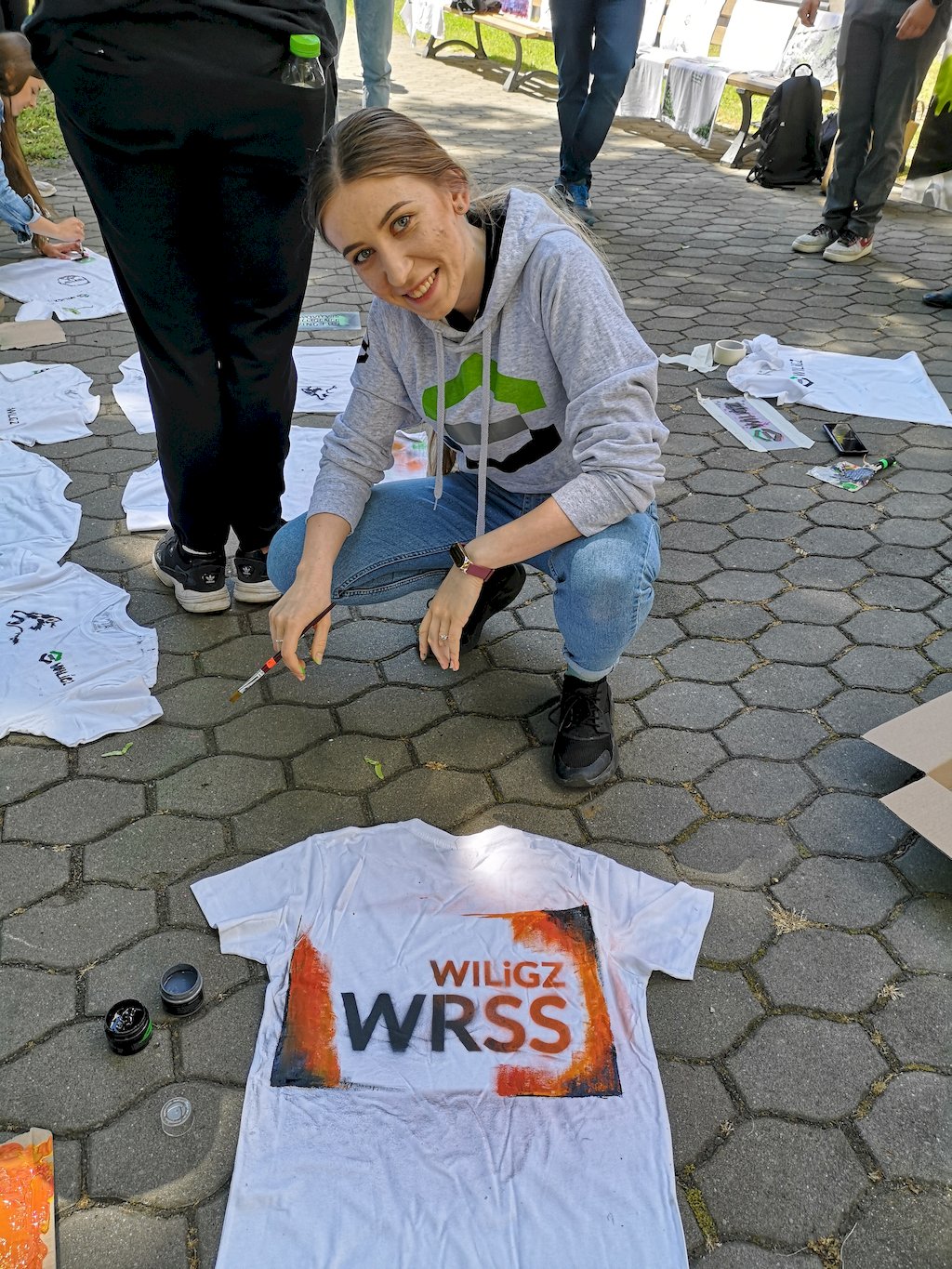 Studentka w szarej bluzie z logo wydziłu i pędzlem w ręce klećzy nad położoną na chodniku z kostki brukowej białej koszulce z napisem WILGZ WRSS