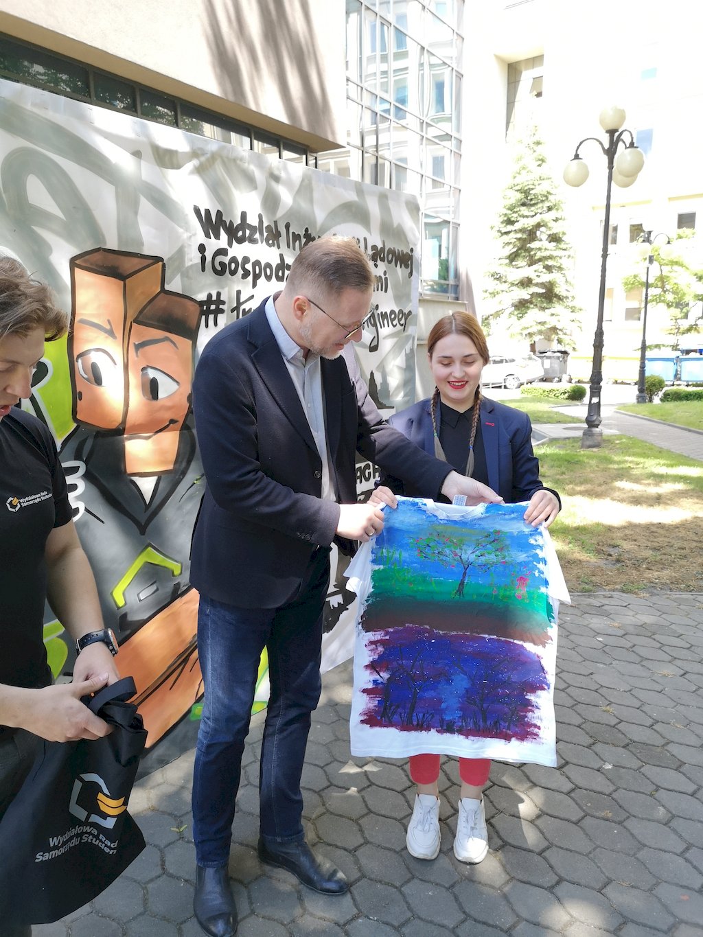 STudentka razem z Dziekanem prof. Markiem Całą prezentują koszulkę z namalowanymi na niej dwoma kolorowymi obrazami prezentującymi drzewo.