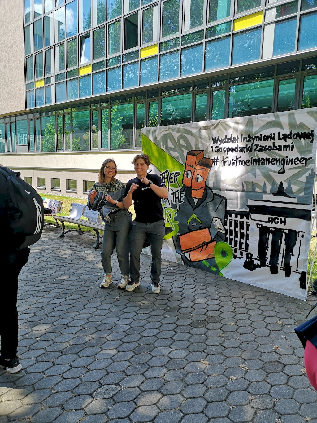Studentka i student tańczą stojąc obok siebie stykając się biodrami na tle plakatu z nazwą wydziłu i konturem budynku głównego AGH