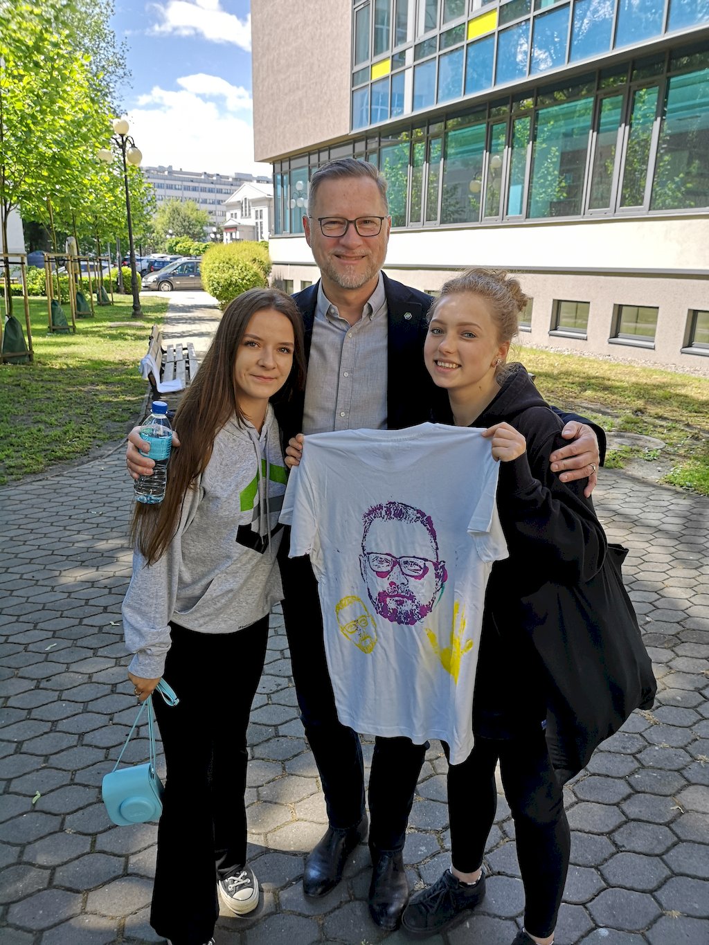 Dziekan prof. Marek Cała stoi pomiędzy dwoma studentkami trzymającymi rozłożoną białą koszulkę z namalowaną twarzą brodatego mężczyzny w okularach.