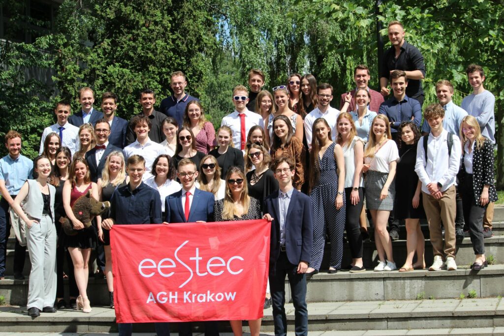 Studenci i studentki stoją na schodach obok siebie. W tle widać drzewa. Studenci na przedzie trzymają czerwoną flagę z napisem: EESTEC AGH Kraków.