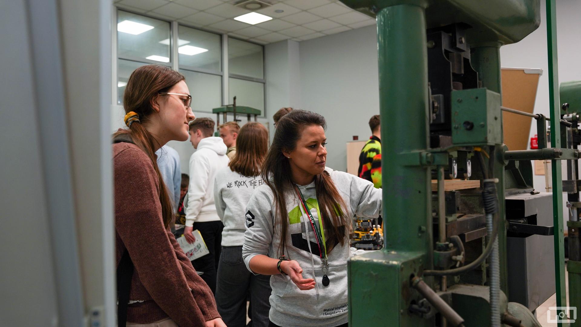 Na zdjęciu widać pracowniczkę Wydziału Inżynierii Lądowej i Gospodarki Zasobami ubraną w szara bluzę wydziałową. Kobieta wyjaśnia młodym ludziom działanie urządzenia. Po prawej stronie kobiety znajduje się maszyna wytrzymałościowa.