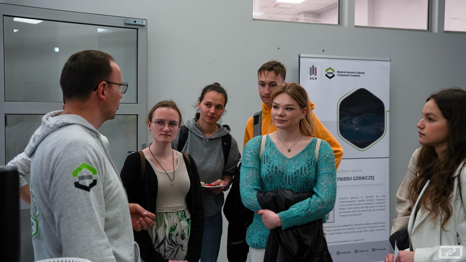 Na zdjęciu widać pracownika wydziału ubranego w szarą bluzę z logiem WILiGZ który rozmawia z młodymi kobietami i mężczyznami. Osoby znajdują się w budynku laboratoryjnym.