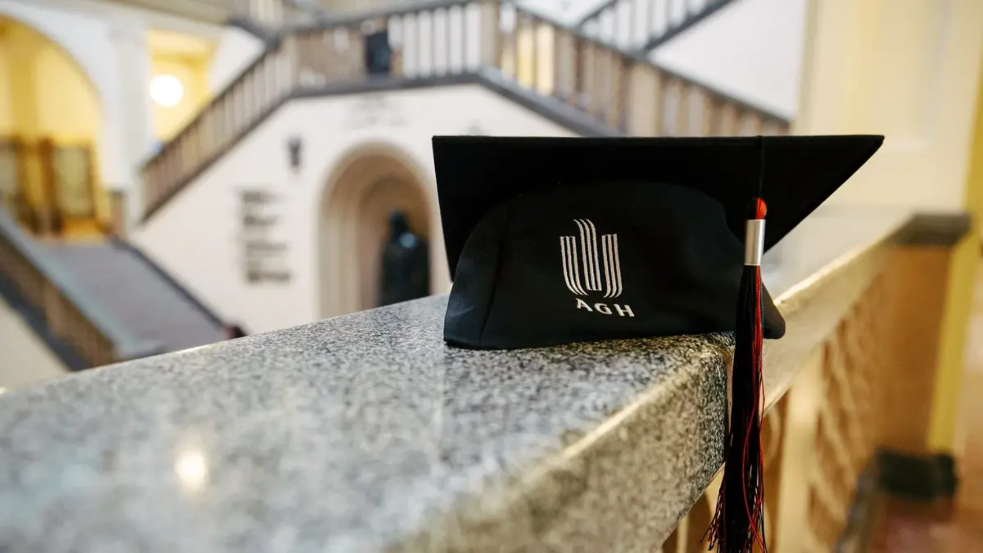 Na zdjęciu widoczna jest czarna czapka absolwenta z logo AGH. Czapka leży na murku w budynku A0.