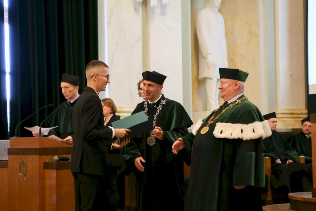 Student odbiera zieloną teczkę od Rektor AGH – prof. dr hab. inż. Jerzego Lisa oraz Dziekana Wydziału Inżynierii Lądowej i Gospodarki Zasobami Dr hab. inż. Arkadiusza Kustry, prof. Uczelni. Prodziekan Wydziału Inżynierii Lądowej i Gospodarki Zasobami ds. Kształcenia dr inż. Romuald Ogrodnik przy mównicy. Osoby na zdjęciu się uśmiechają.